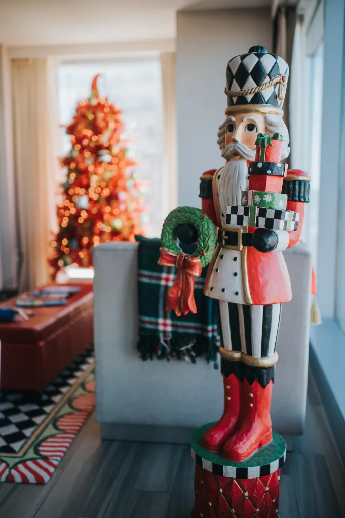 A nutcracker statue wearing red boots and a green wreath, holding colorful gift boxes, stands in a room with a Christmas tree.
