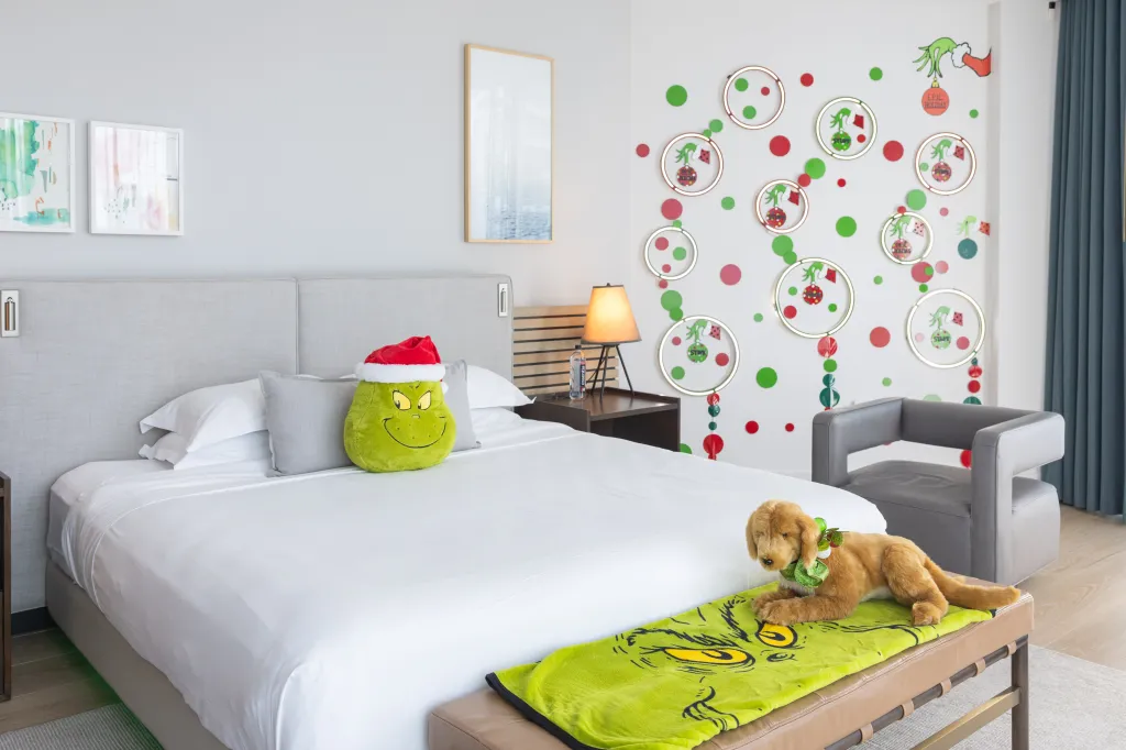 A bedroom with a Grinch pillow, blanket, and wall decorations, featuring a plush dog with a holiday collar.