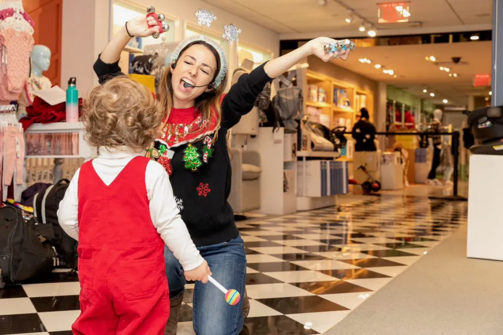 An adult woman in a festive sweater and snowflake headband leads a child in red overalls in a music activity.