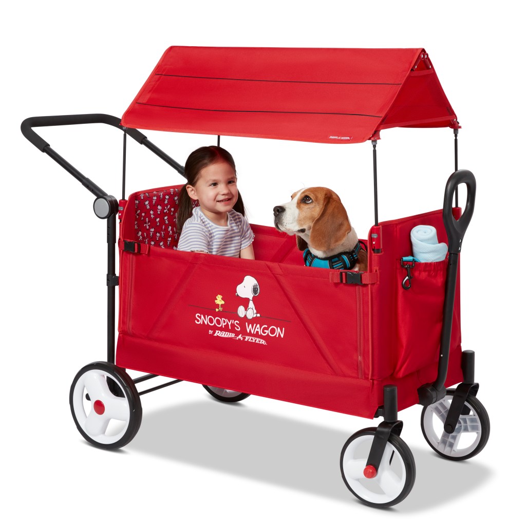 A little girl and a beagle inside a Radio Flyer Snoopy's Wagon with a canopy.