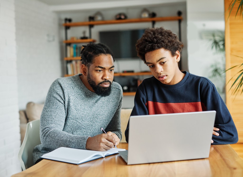 Father and teenage son doing homework together using a laptop at home.