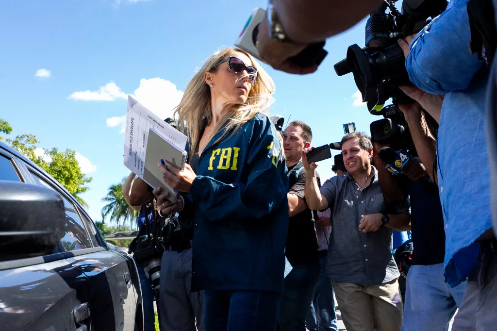 An FBI agent in a blue jacket leaving an Autozone while holding papers and being filmed by multiple news cameras.