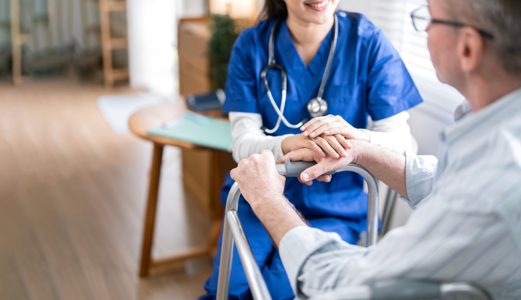 A female doctor in blue scrubs and a stethoscope holding the hand of an older male patient with a walker.