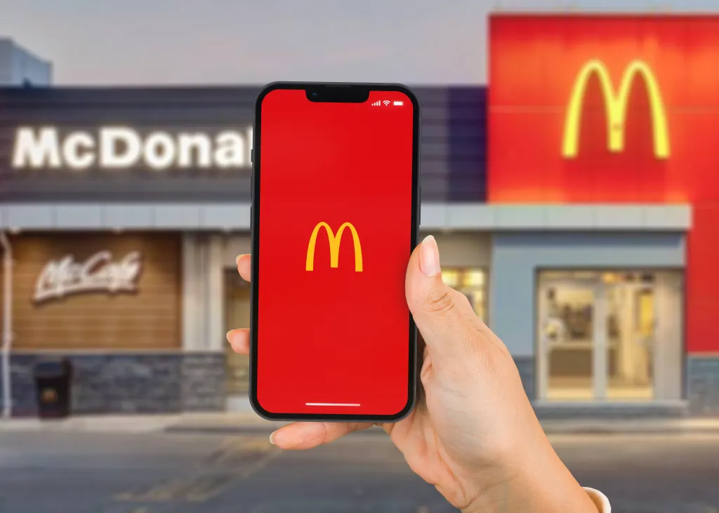 Hand holding a smartphone displaying the McDonald's app with a blurred McDonald's store in the background.
