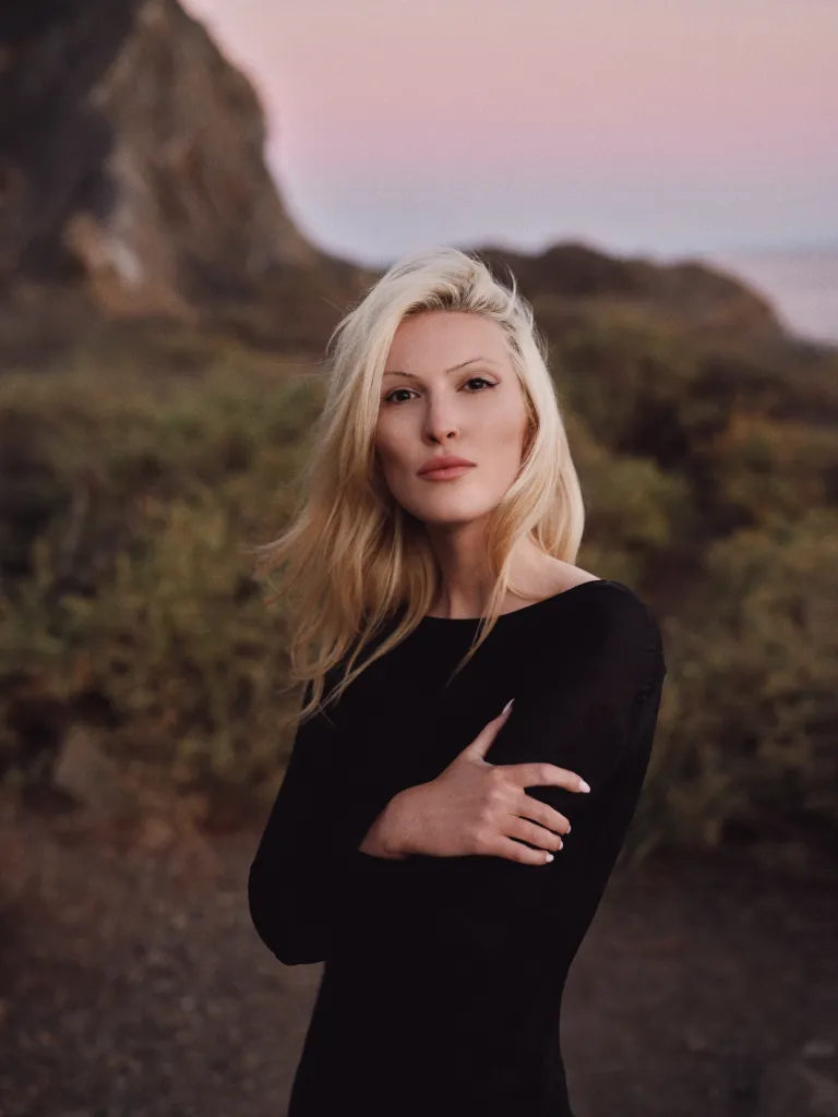 Olivia Nuzzi in a black dress with her arms crossed, standing outdoors with a hillside and pink sky in the background.