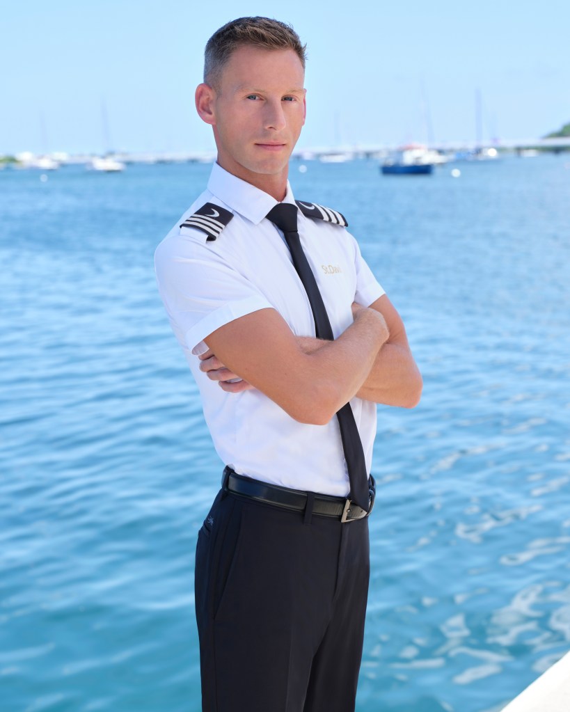 Fraser Olender from Below Deck Season 12, wearing a white uniform shirt with gold 'St. David' embroidery, a black tie, and black pants, standing with crossed arms by the water.