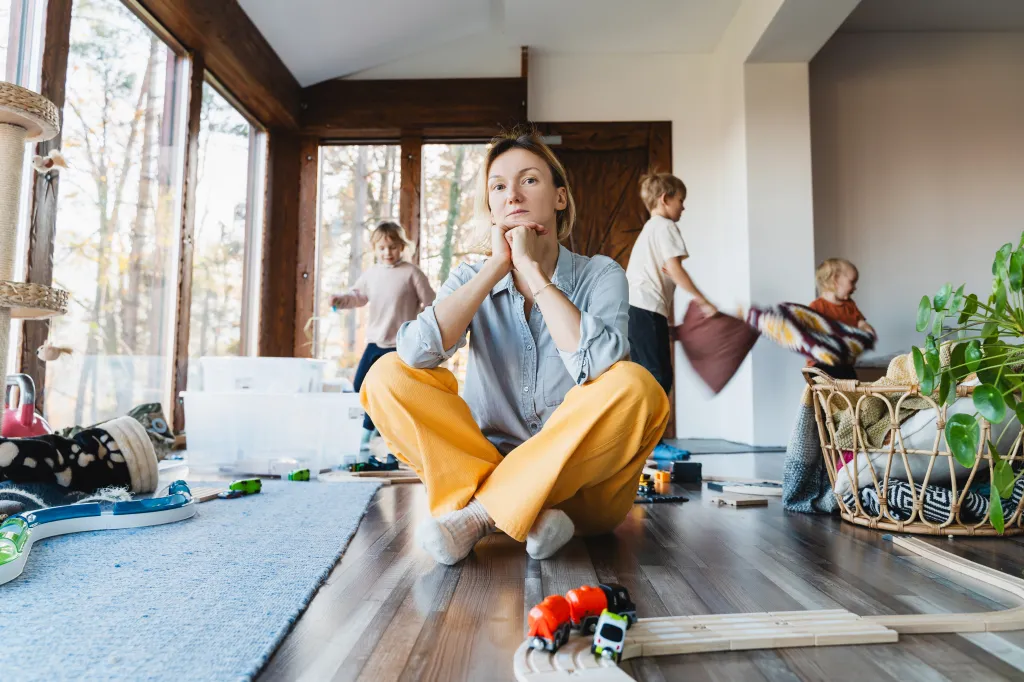 A stressed mother sits on the floor surrounded by toys, while her children play noisily around her.
