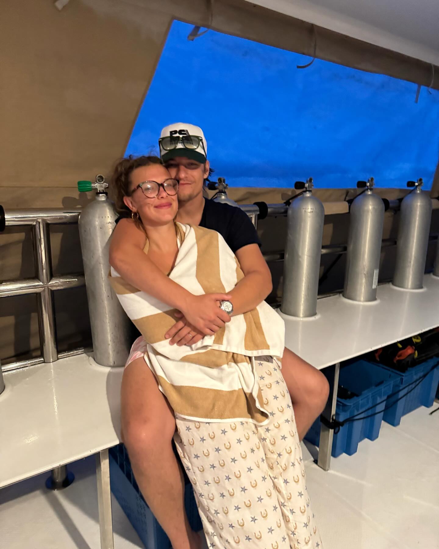 Millie Bobby Brown and Jake Bongiovi embracing on a boat with scuba tanks visible.