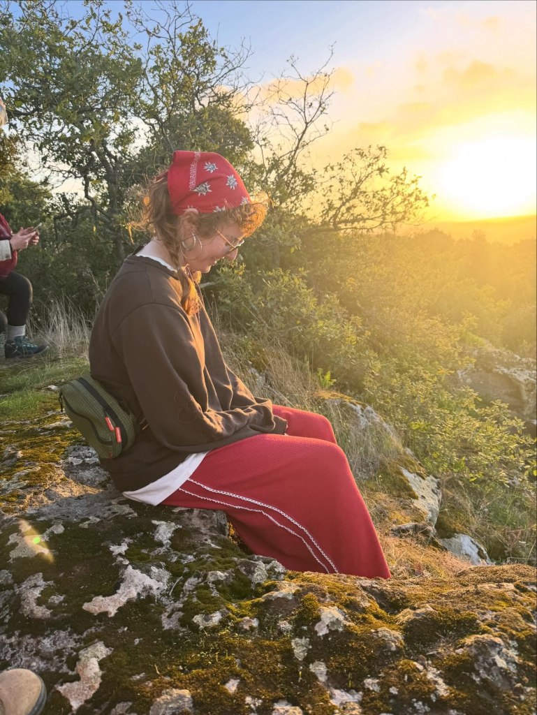 Maisie Williams' friend sitting on a mossy rock in a red bandana and red pants, with a sunset behind her.