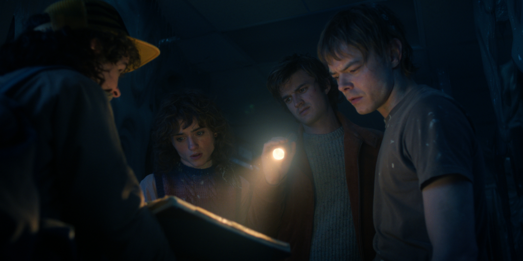 Dustin, Nancy, Steve, and Jonathan look at a book, illuminated by Steve's flashlight.