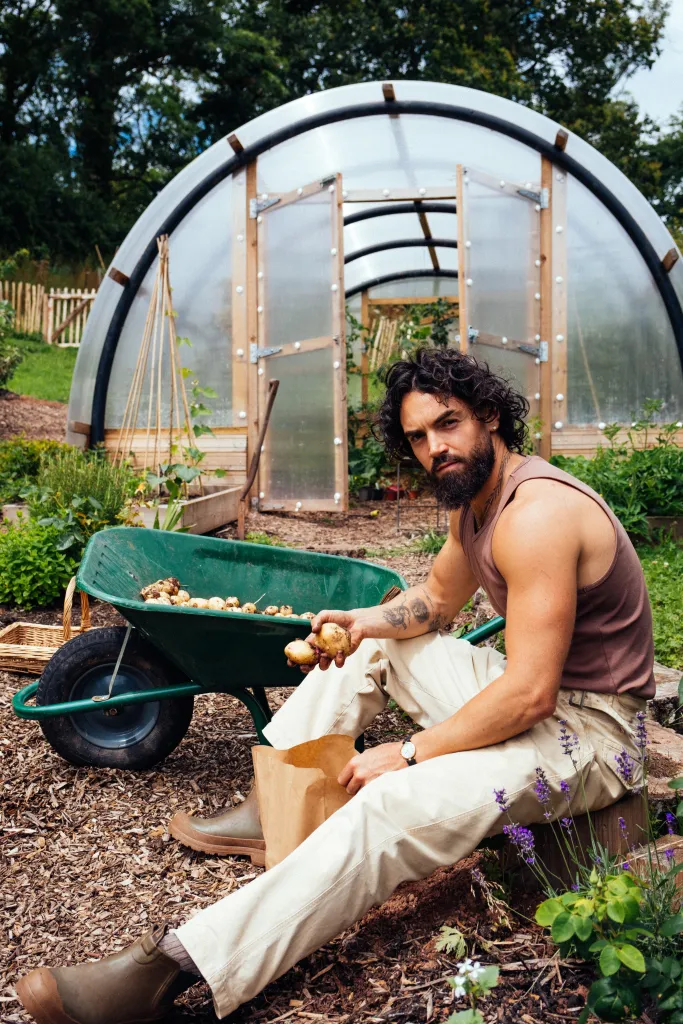 Celebrity chef Gaz Oakley sitting next to a wheelbarrow full of potatoes, holding a potato, with a greenhouse in the background.