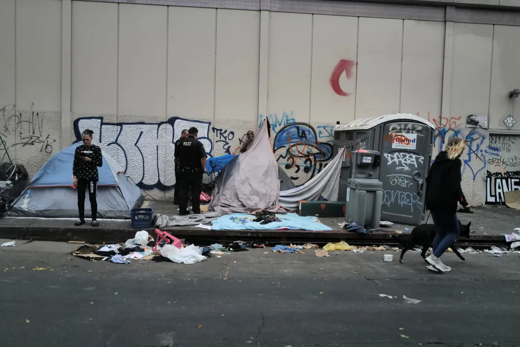 A Skid Row street scene shows tents and a portable toilet along a graffiti-covered wall, with two police officers standing near a tent and a woman walking a black dog past scattered trash.