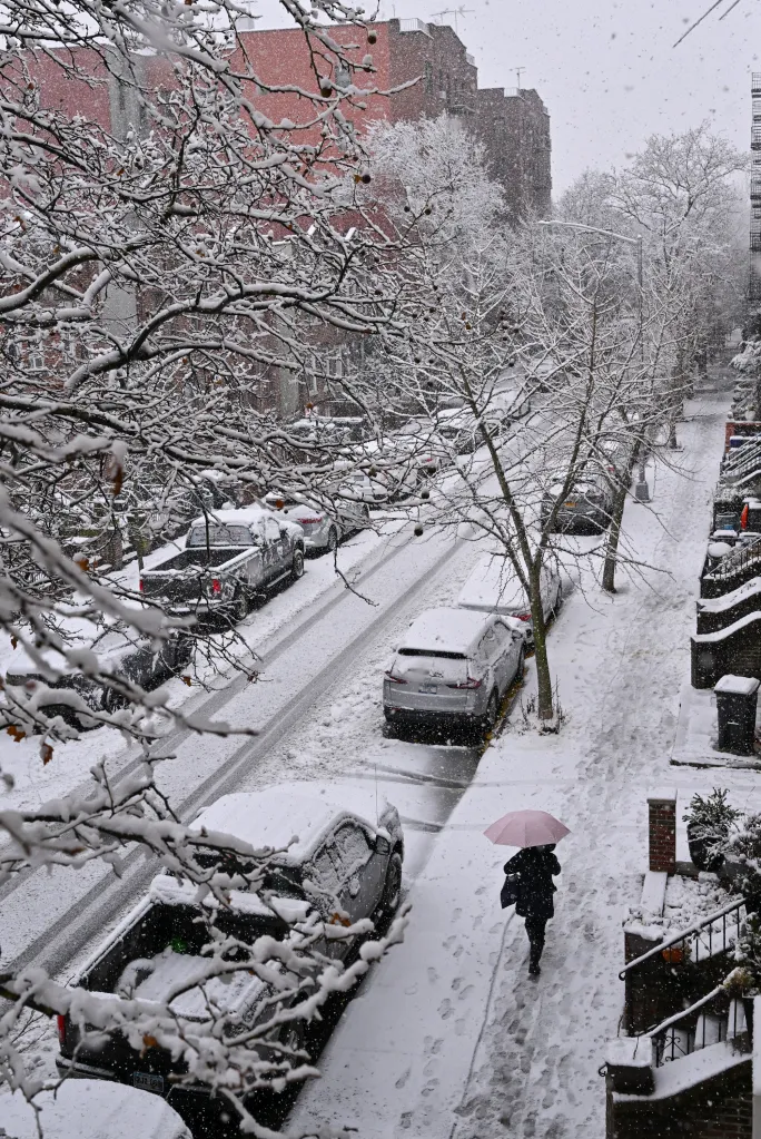 Overhead view of a snowy Brooklyn street with parked cars and a person with a pink umbrella.