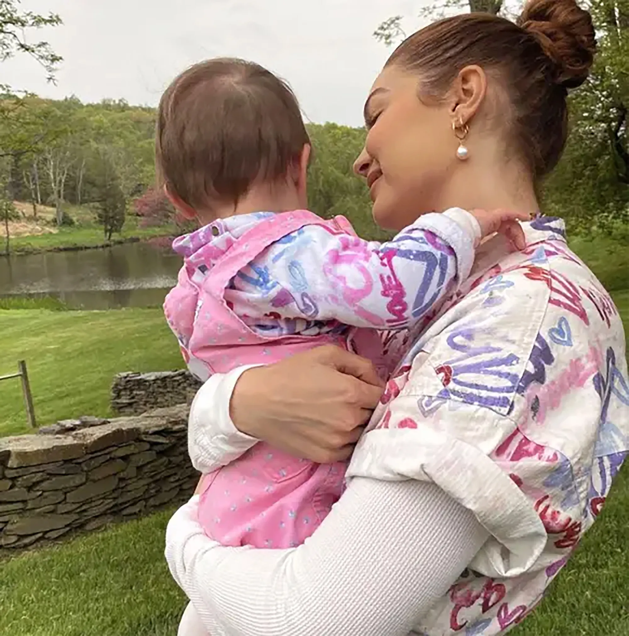 Gigi Hadid holding daughter Khai, who is wearing a pink outfit, outdoors in a scenic area with a lake.