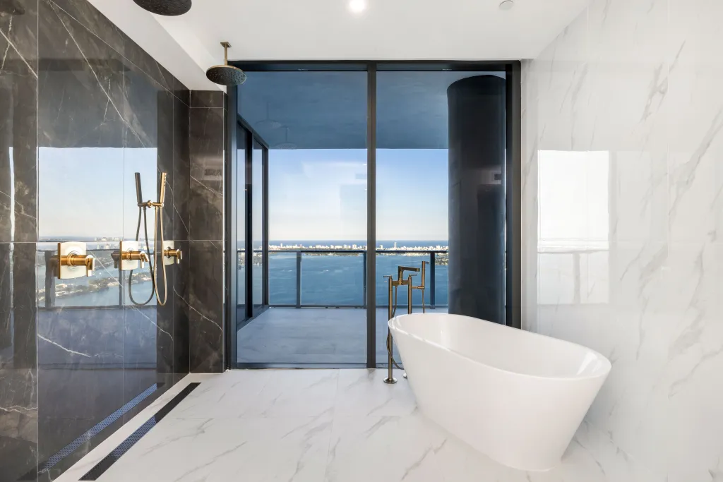 A modern bathroom with a freestanding white bathtub and an open shower area, featuring large windows offering views of Biscayne Bay and the Edgewater District.