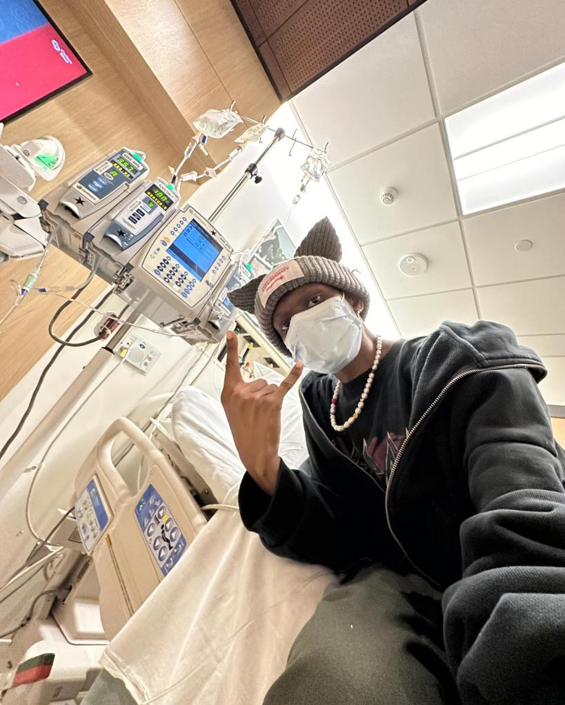 A person in a hospital bed with an IV drip, wearing a face mask and a cat-eared hat, makes a rock-on gesture.