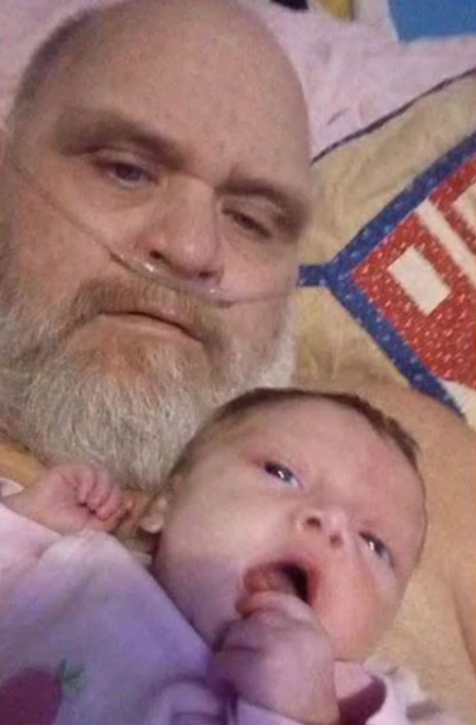 A bald man with a beard and an oxygen tube across his face next to a baby with a hand in its mouth.