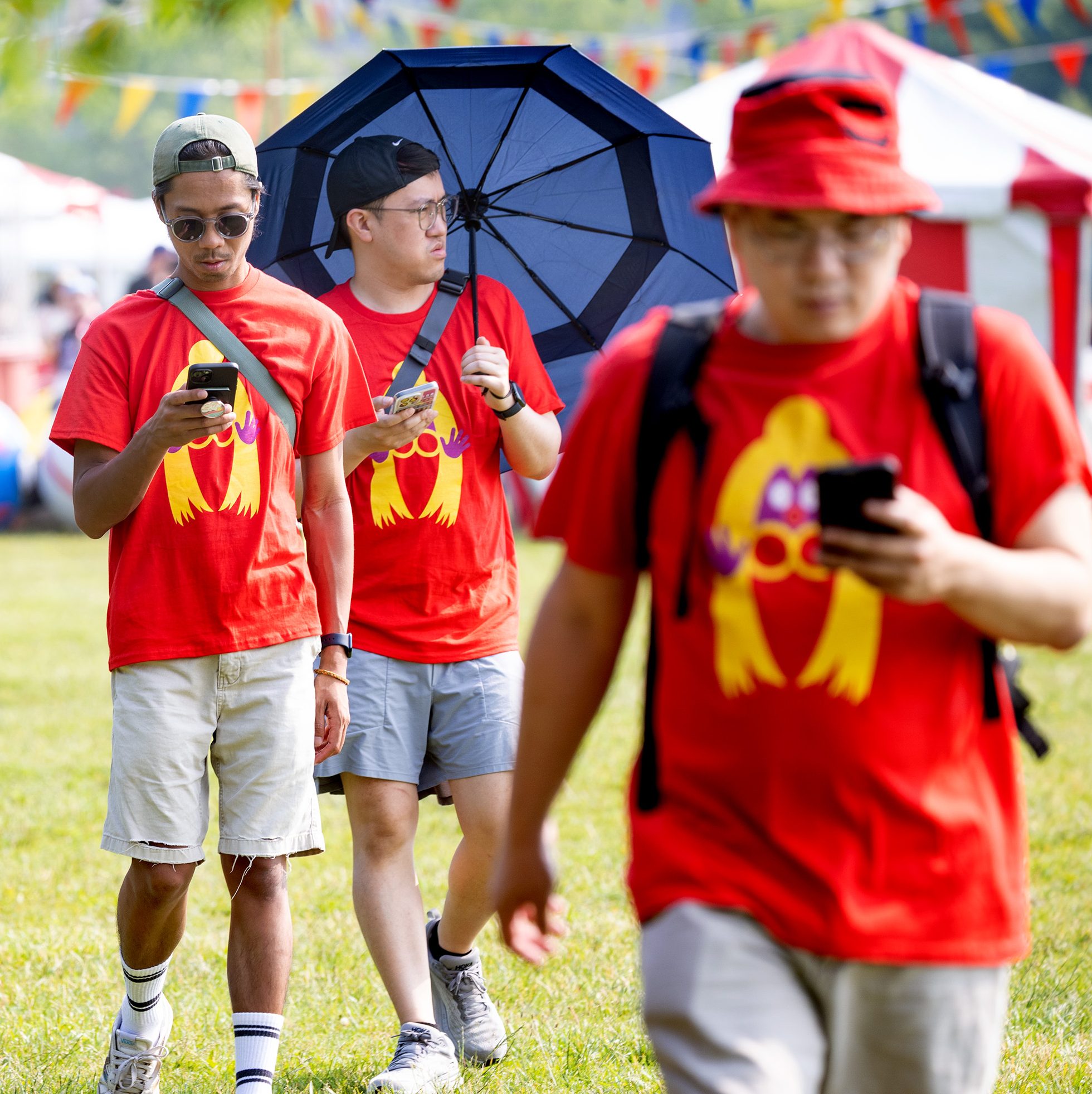 Four Pokémon trainers in red shirts walk across a grassy field at Liberty State Park, two of them holding umbrellas.
