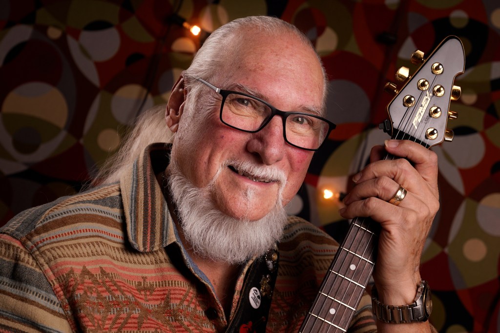Steve Cropper, an older white man with a white beard, long white hair, and black-framed glasses, smiles while holding a black and gold guitar.