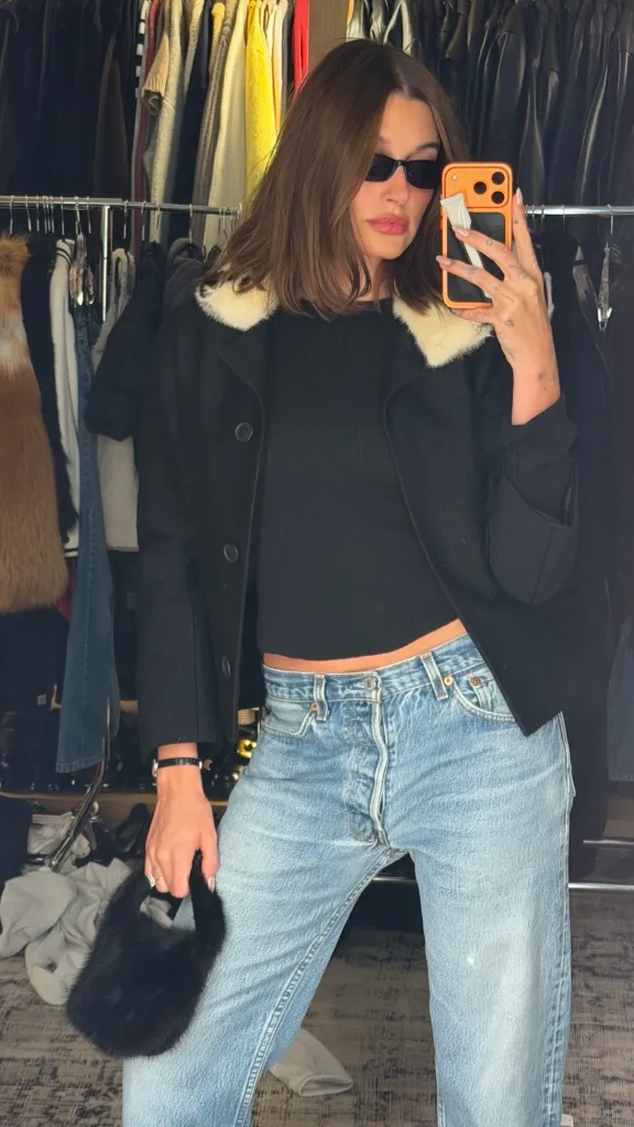 Hailey Bieber taking a mirror selfie wearing a black coat with a white fur collar, jeans, sunglasses, and holding a small black fur handbag.