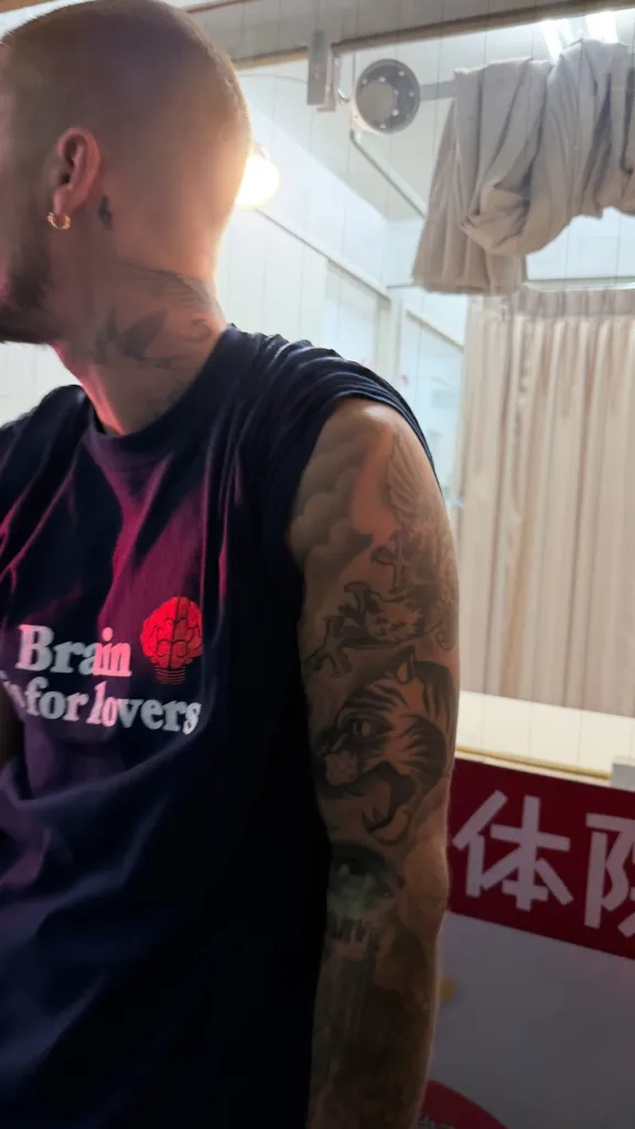 Person with buzzcut, gold hoop earring, and tattoos on neck and arm, wearing a 