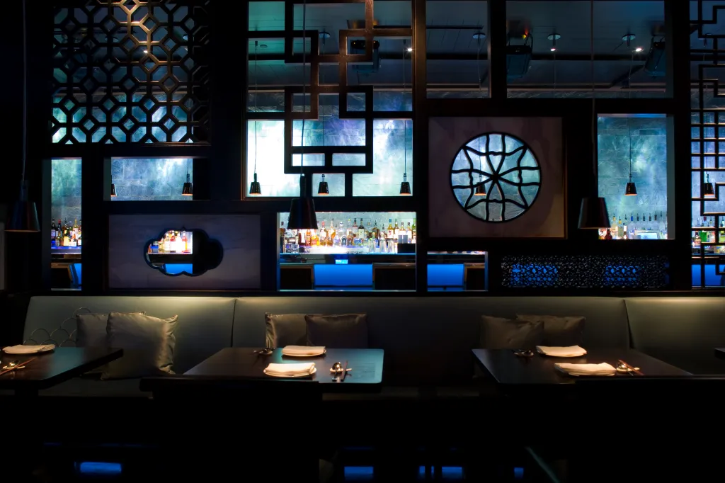 Interior of a dark, stylish restaurant with blue backlighting behind the bar and decorative screens.