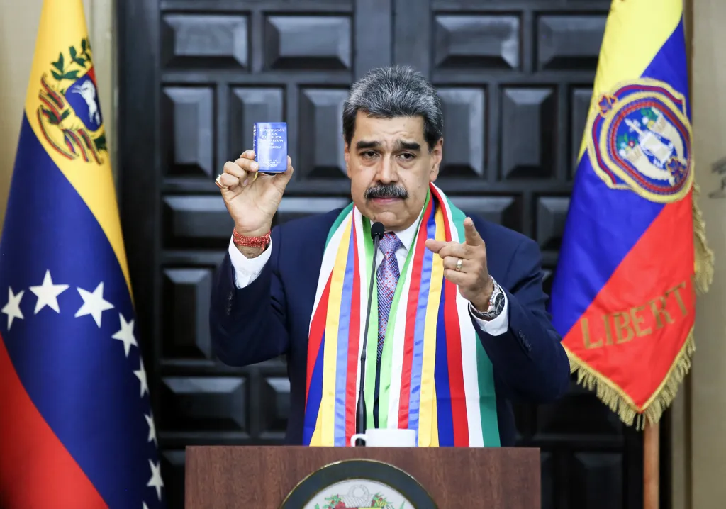 Venezuelan President Nicolas Maduro holding up a blue book and pointing with his right hand.