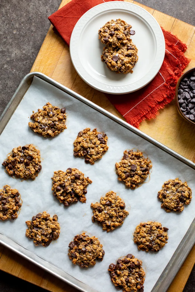 Freshly baked oatmeal chocolate chip cookies, some on a baking sheet and two on a white plate with a red napkin.