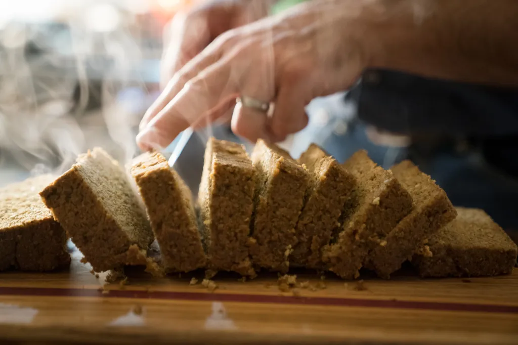 A person slicing a loaf of bread, with steam rising from the warm slices.