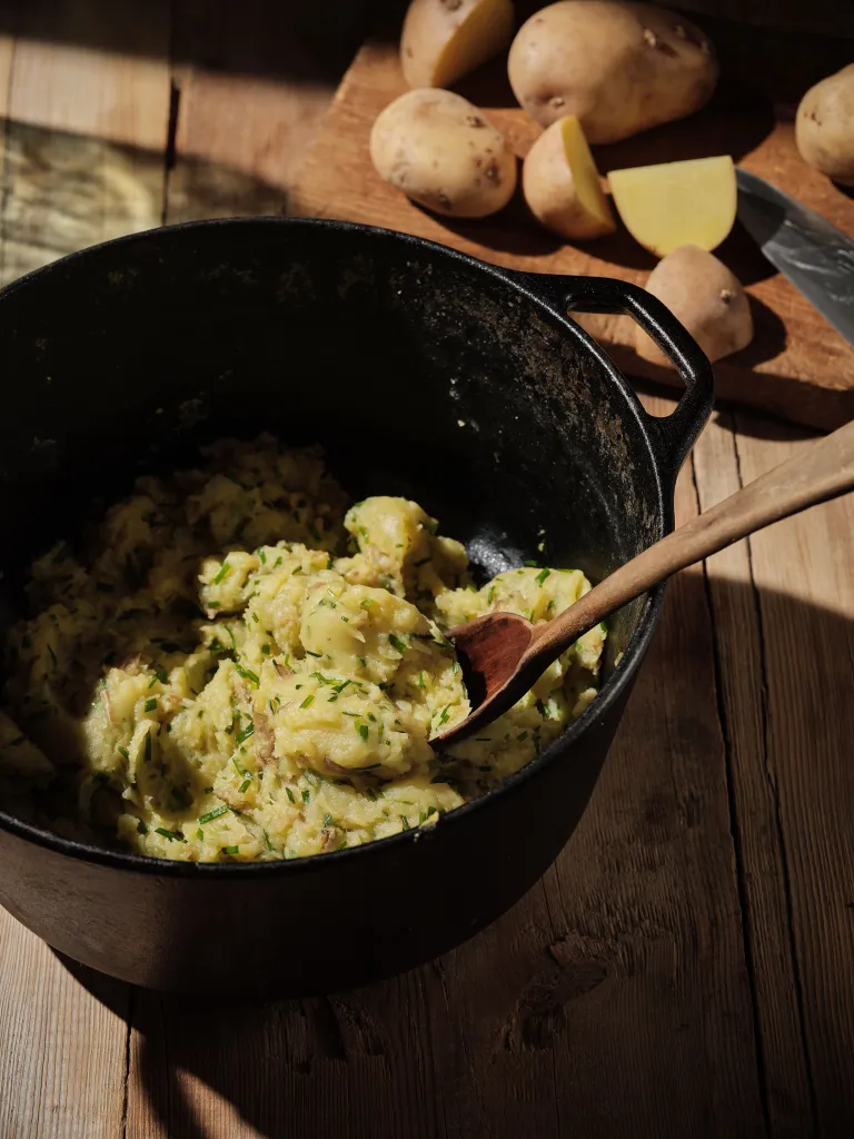 Mashed potatoes with chives in a cast iron pot next to whole and cut potatoes and a knife on a wooden cutting board.