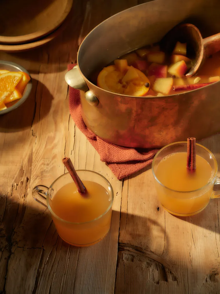 Two mugs of cider with cinnamon sticks next to a copper pot of cider, fruit, and spices.