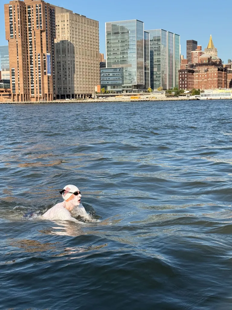 Michael Moreau with a white swim cap and dark goggles swimming in a body of water with city buildings in the background.