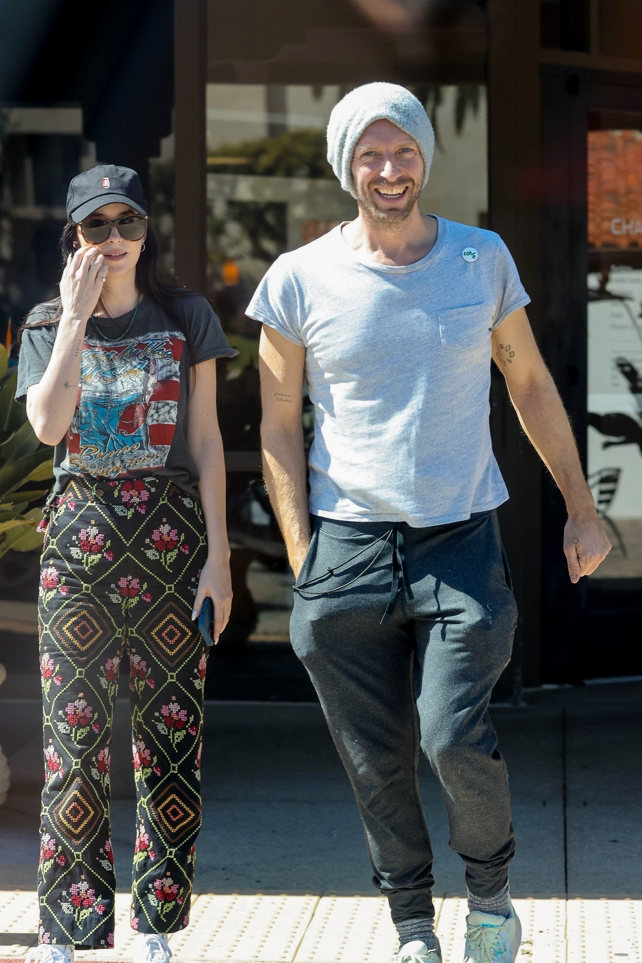 Chris Martin and Dakota Johnson walking with Martin smiling, wearing a light gray beanie and shirt with a 
