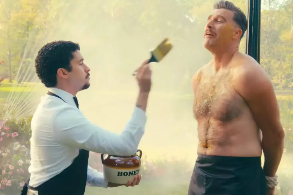 Travis Kelce tied to a post while a man applies honey to his chest and face with a brush.