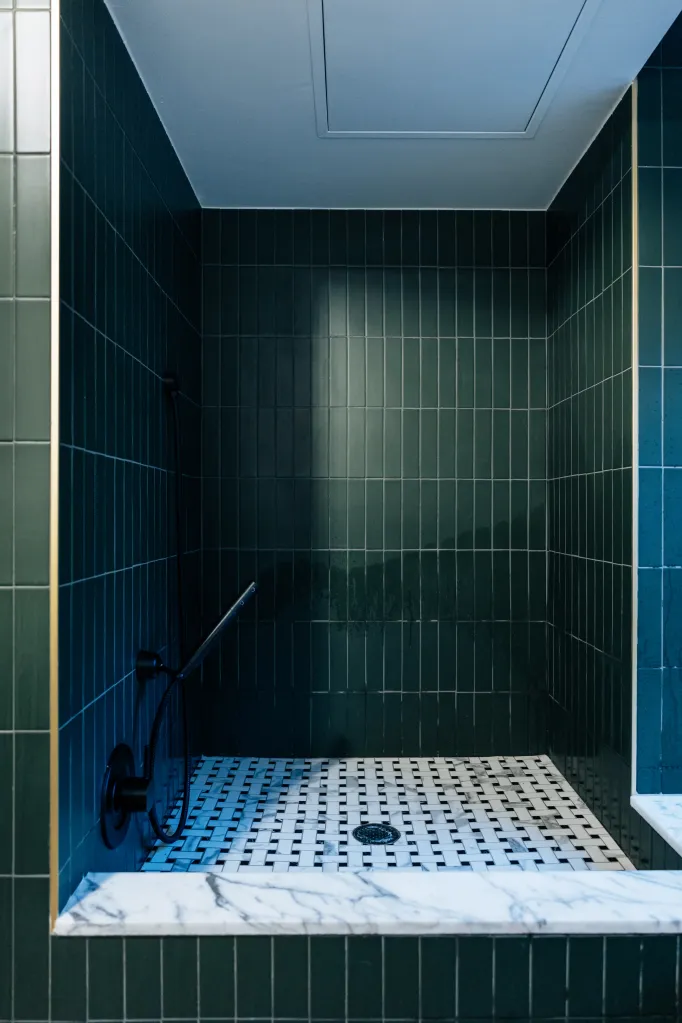 A shower stall with dark green rectangular tiles, a white and black mosaic floor, and a black showerhead.