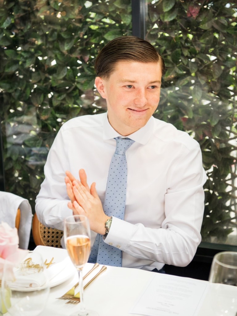 18-year-old Henry Nowak in a white shirt and blue tie, clapping his hands at a table with a glass of rose champagne.