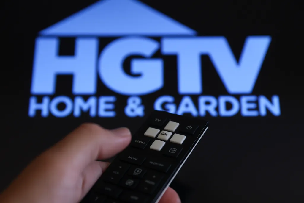 A hand holding a remote control in front of a TV screen displaying the blue HGTV logo and the words 