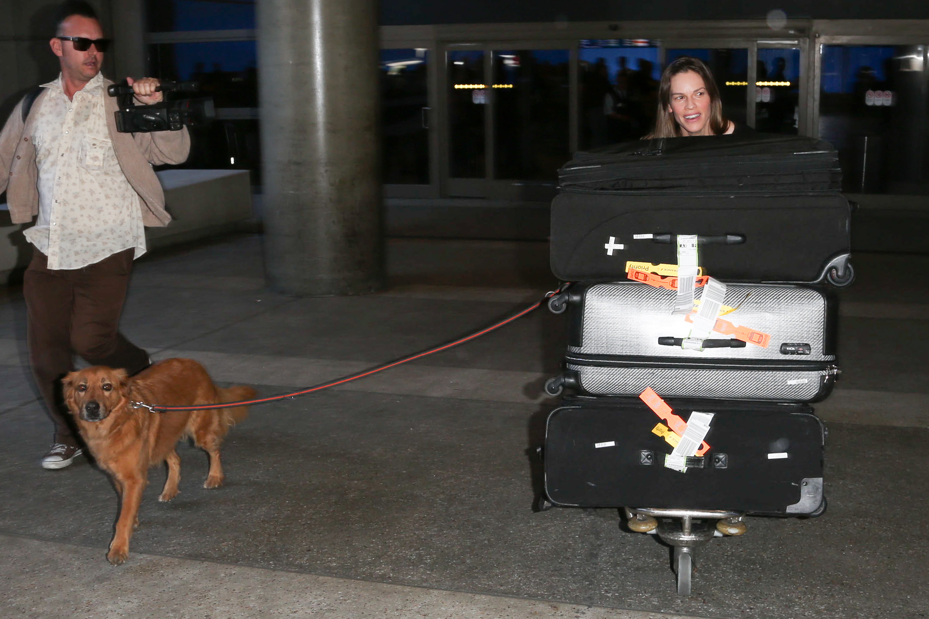 Hilary Swank at LAX Airport with a man, a dog, and several pieces of luggage on a cart.