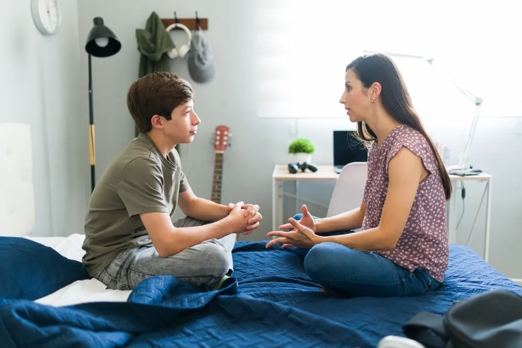Hispanic mother and teenage son having a serious conversation in his bedroom, both sitting on the bed.