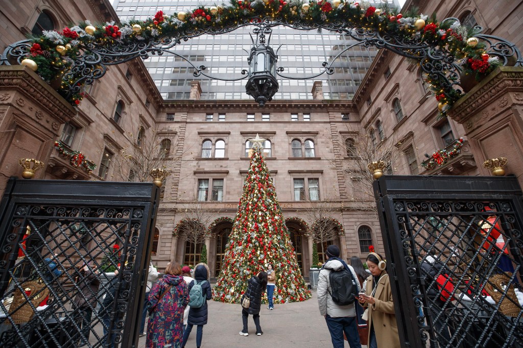 A large Christmas tree decorated with red and gold ornaments stands in the courtyard of Lotte New York Palace Hotel, with a decorated archway in the foreground.