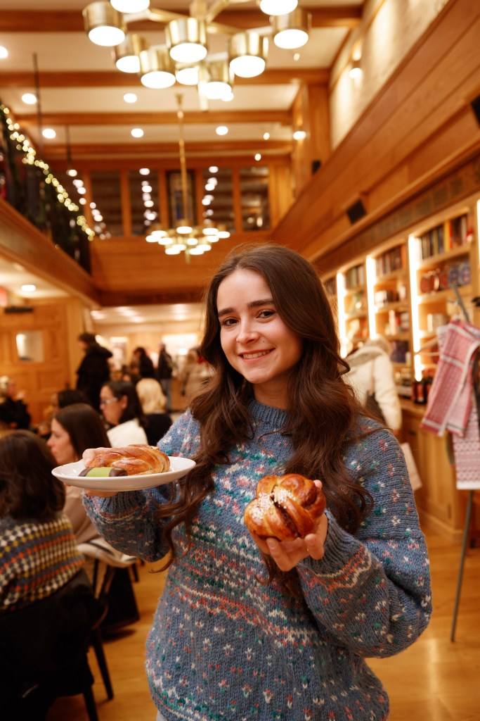 A smiling young woman in a blue patterned sweater holds a plate with a Swedish cinnamon bun and a marzipan pastry in one hand and another cinnamon bun in the other, while standing in a gift shop.