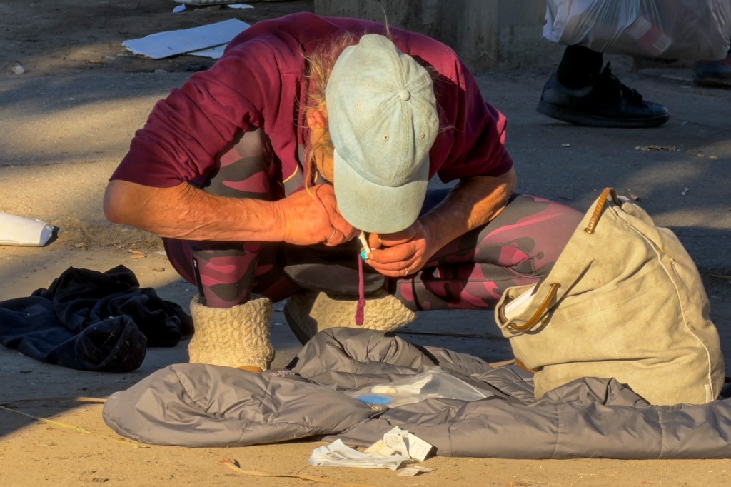 A person in a baseball cap crouches on the ground, preparing to inject a substance.