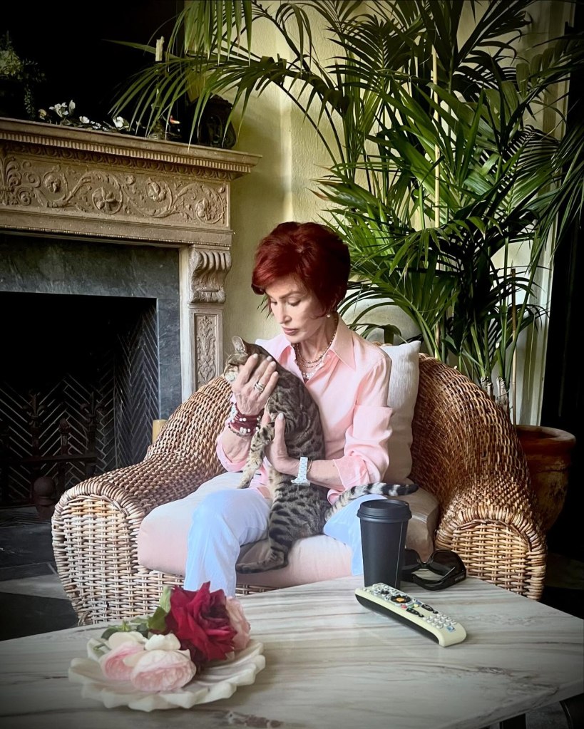 Sharon Osbourne sitting in a wicker chair, holding a cat, with another cat in her lap.