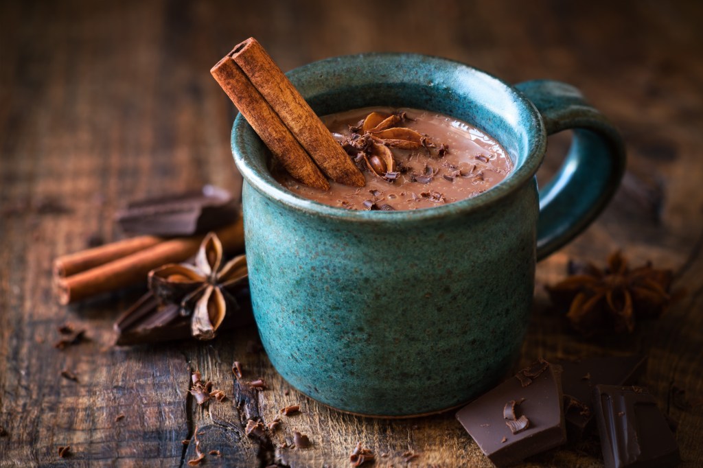 A mug of hot chocolate with a cinnamon stick, star anise, and grated chocolate on a wooden surface.