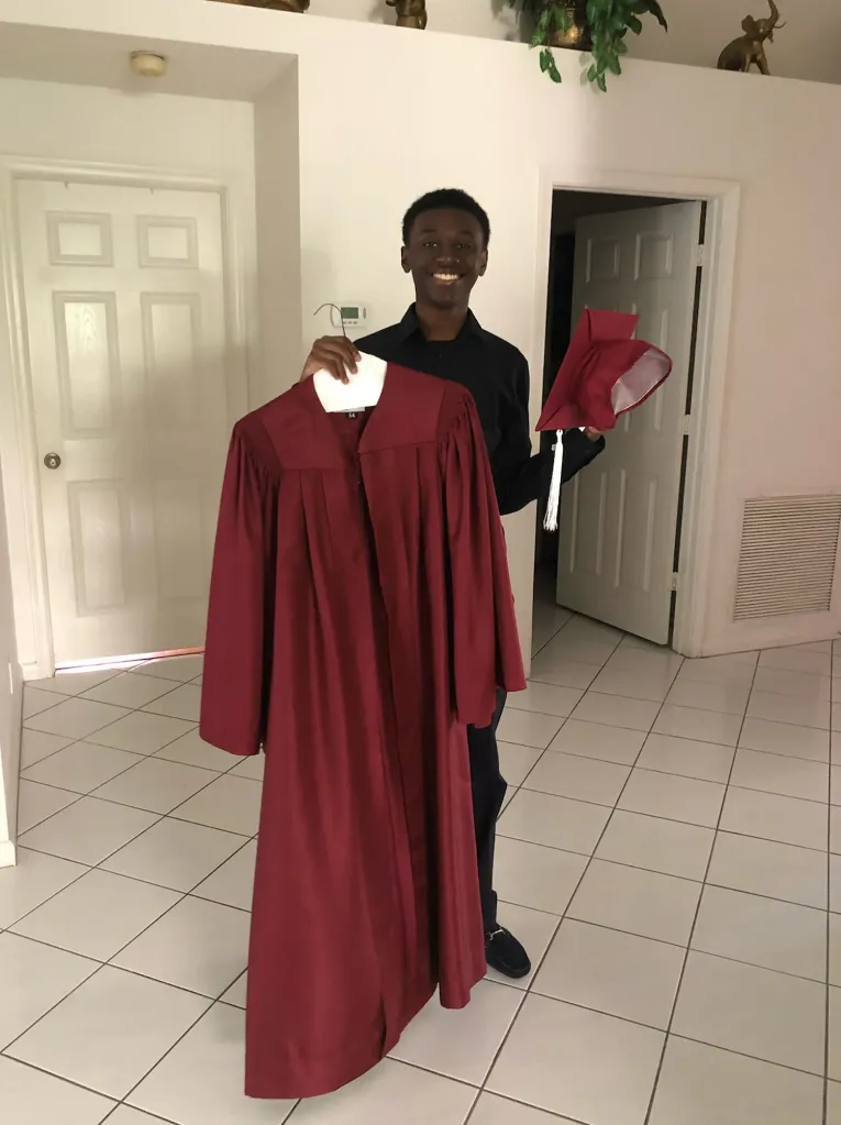 A young man smiles while holding a burgundy graduation gown and cap.