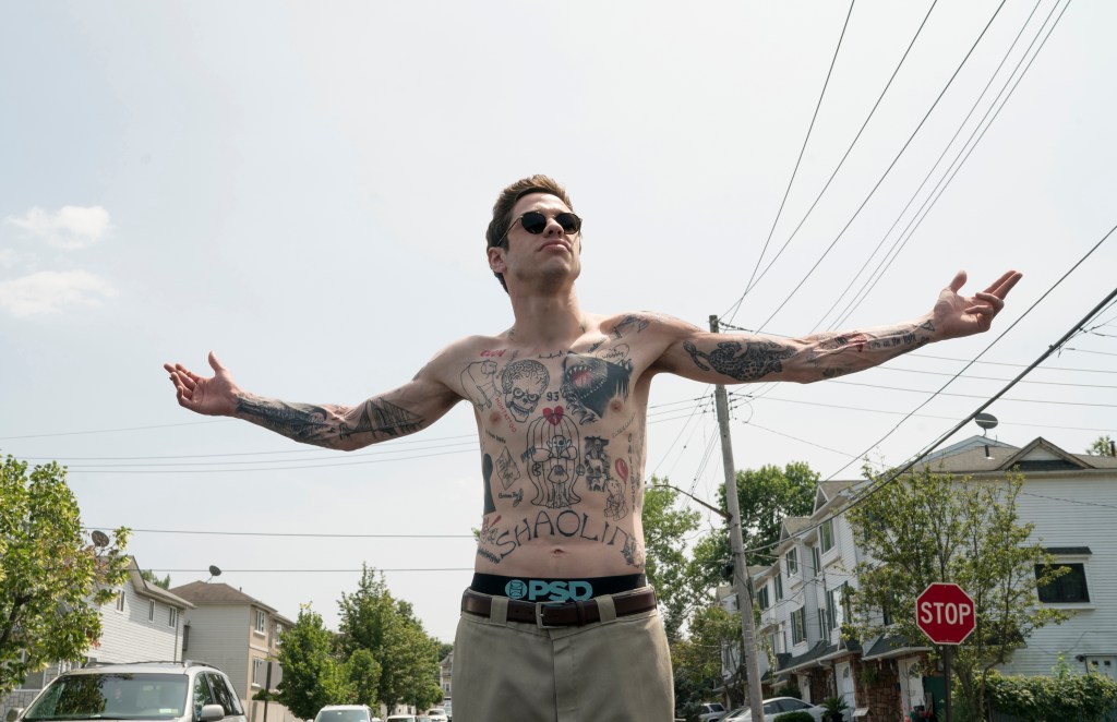 Pete Davidson with his shirt off and arms outstretched in a residential area.