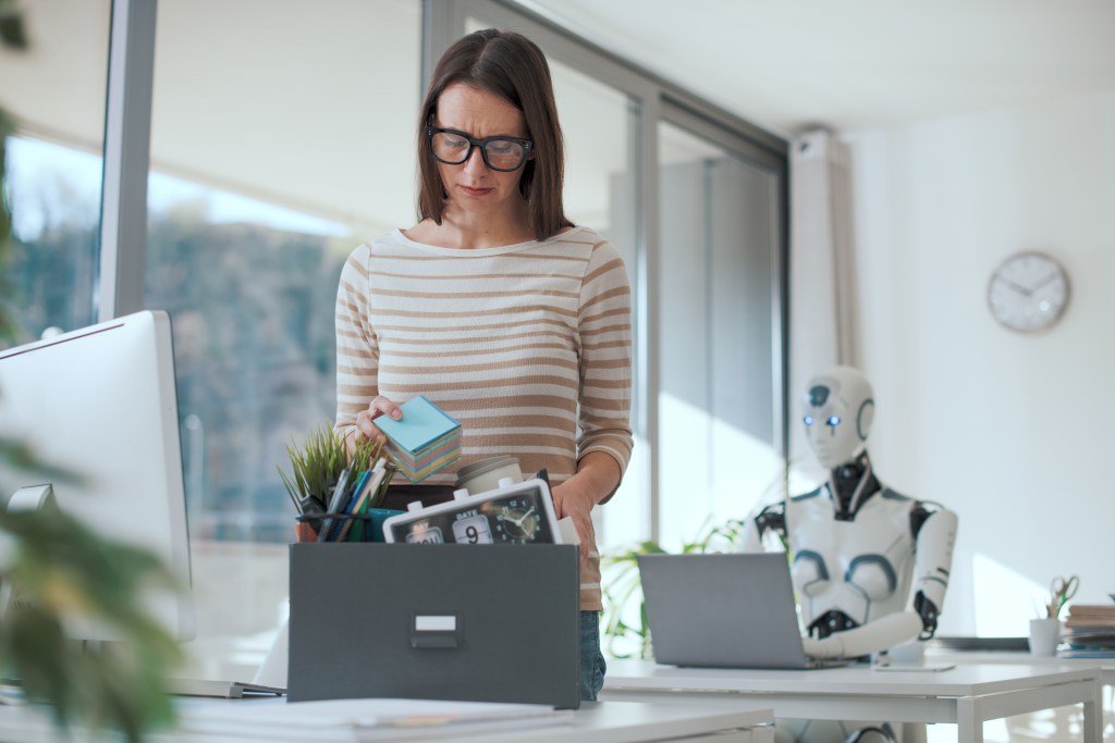 Woman packing her belongings while a robot works at a desk, illustrating AI's impact on employment.