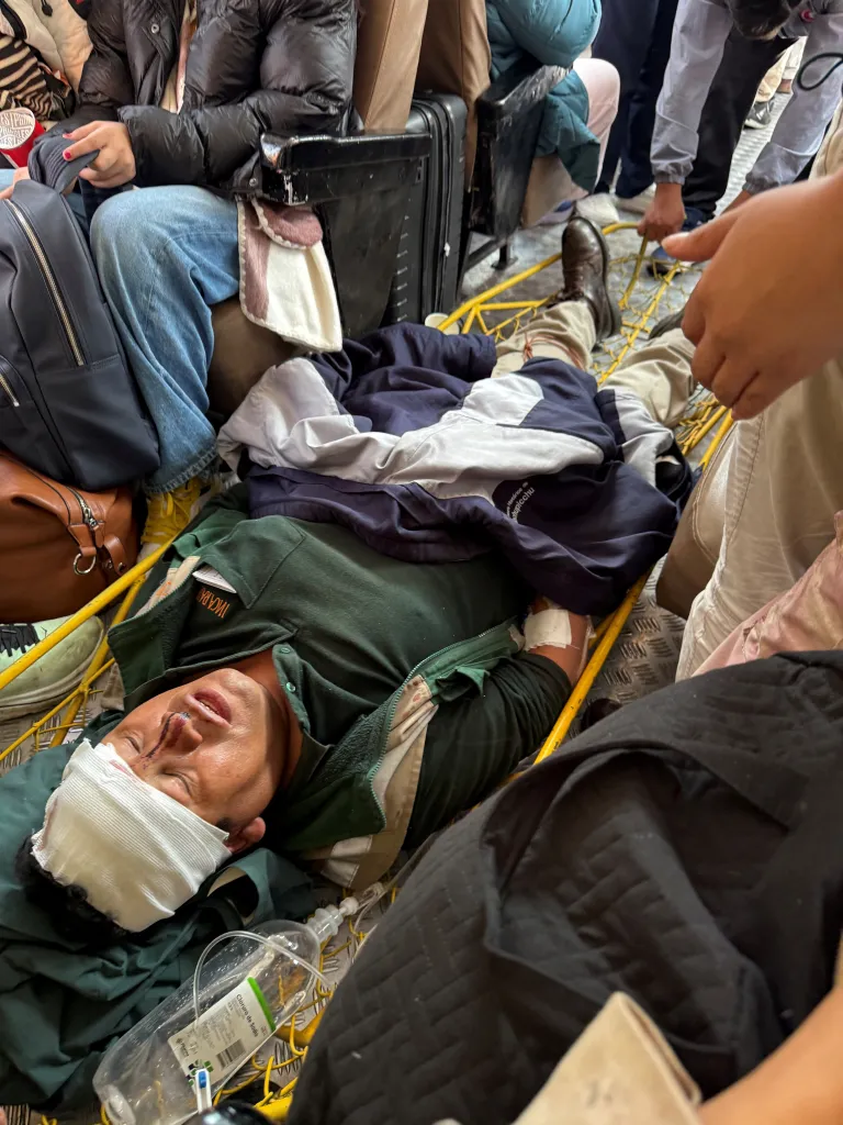 An injured person with a bandaged head and blood on their nose lies on a stretcher, with an IV bag nearby.