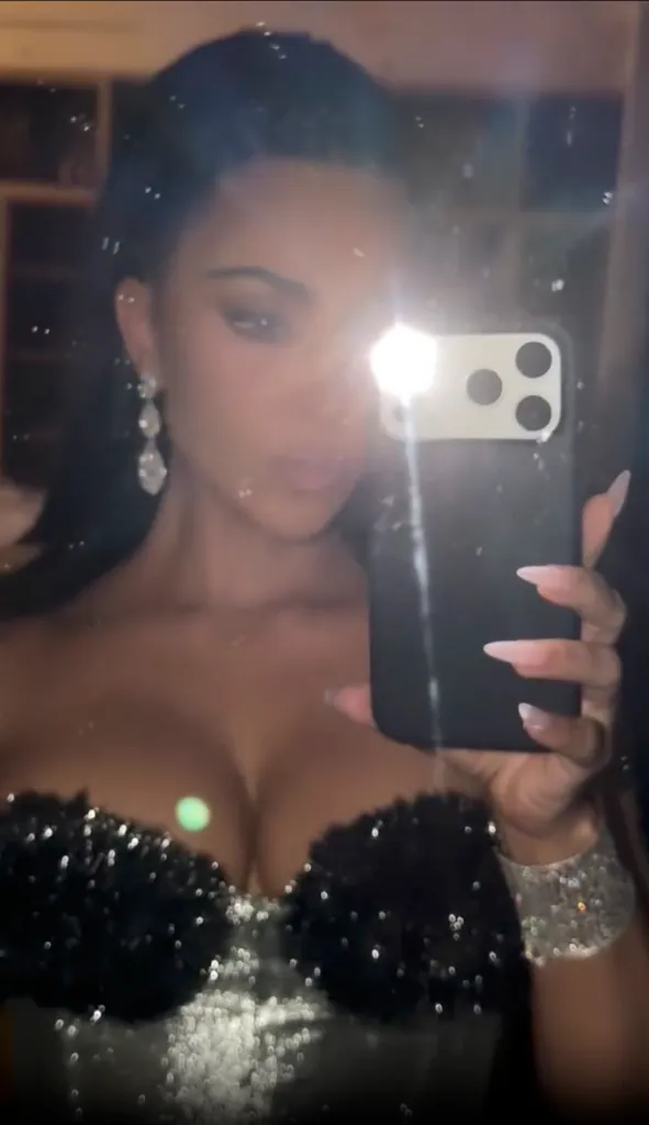 Kim Kardashian taking a mirror selfie, dressed in a black glittery top, wearing long earrings and a sparkly bracelet.