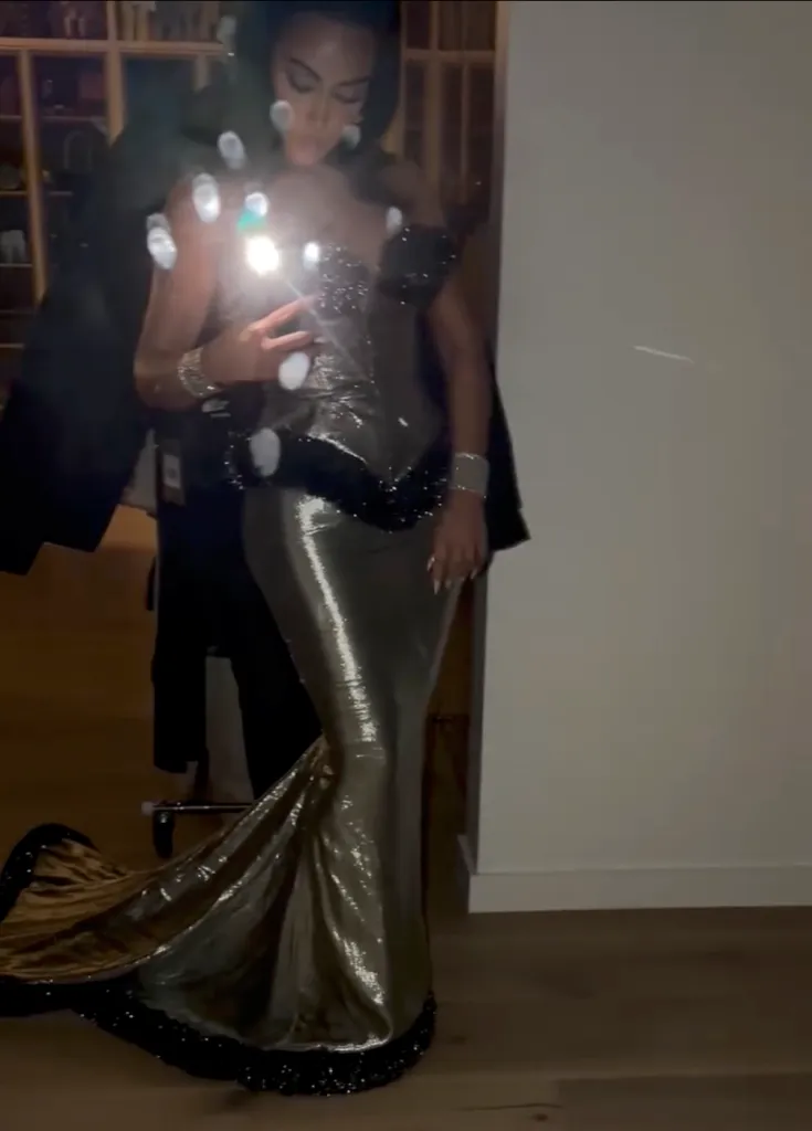 A woman in a gold sequined gown with a black feather detail at the waist holding a phone with a bright flash.